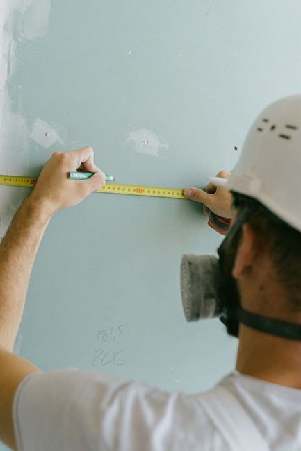 A worker wearing a white safety helmet and a protective mask is measuring the distance between two marks on a light blue painted wall using a yellow measuring tape. The worker's hands are visible, hol